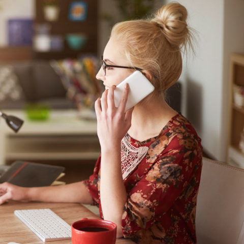 blonde bespectacled woman seated at desk speaking on phone