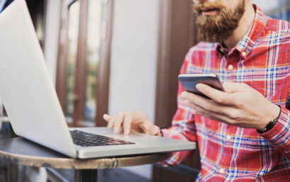 Man sits at a laptop with a computer, holding on to his phone as if he's logging in on both devices.