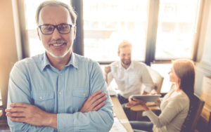 Good Reasons to Retire Later Older Man standing with arms crossed and smiling with two others sitting behind him.