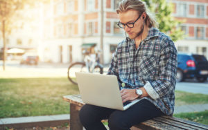 College Student outside class on a bench working on his laptop with ear buds in.