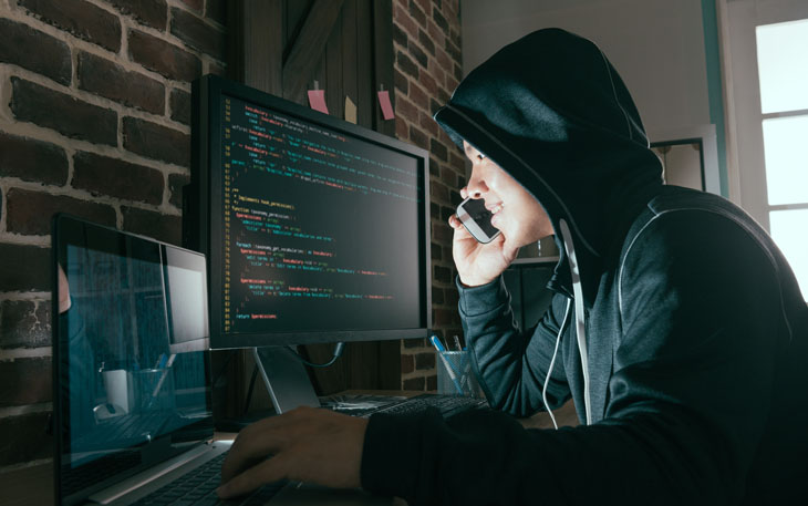 Man in black hooded sweatshirt sits at a desk with two computers on the phone in a dark room