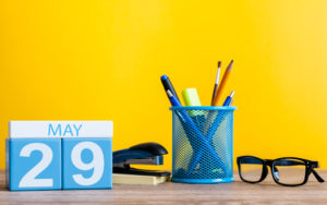 Desk with yellow background. Blocks feature the words May 29 are shows with a stapler, cup with pencils and pens, and a pair of glasses.