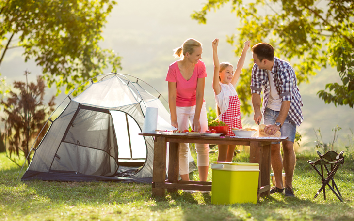 Family with young daughter stand outside their tent in the sunshine. A cooler is in front of them and they're all smiling, enjoying camping.