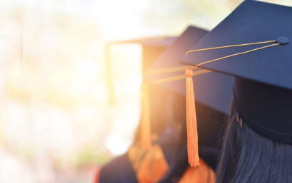 Photo of three high school seniors. Picture focuses on the backs of their heads, showing their graduation cap and tassel. The sun is in shining in front of them.