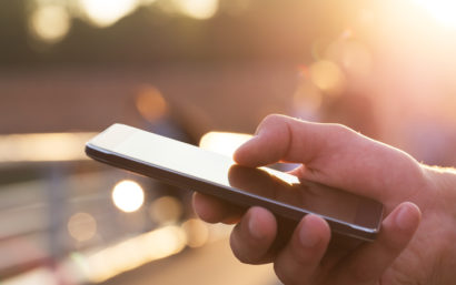 A man's hand holds a smartphone. The background is sunny.