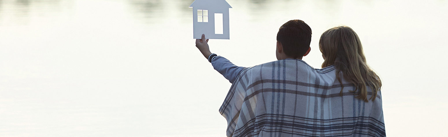 Couple wrapped in a blanket holding up a cut out of a home so one day they will have a mortgage