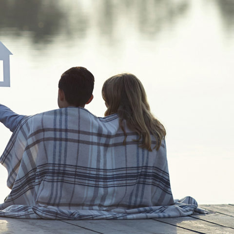 Couple wrapped in a blanket holding up a cut out of a home so one day they will have a mortgage