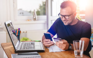 Man sits at a table with a laptop in front of him using his phone to type in a credit card number.