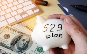 Hand holding a piggy bank with "529 plan" written on it. A few hundred dollar bills lay on a table.