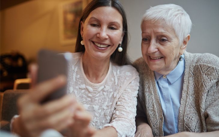 Adult daughter and elder mother smile while looking at a phone.