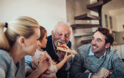 Adult children, grandchild and grandfather eating pizza and smiling.